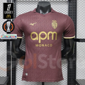 AS Monaco burgundy short-sleeve jersey with tonal pattern, gold crest and APM Monaco sponsor, front view alongside Ligue 1 and Europa League graphics.
