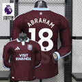 Front and back views of a claret Aston Villa long-sleeve jersey with a light blue collar, white Abraham number 18 print and Visit Rwanda sponsor, alongside Premier League graphics.