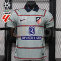 Front view of a light grey Atlético Madrid short-sleeve jersey with red horizontal stripes, a navy collar, club crest and Riyadh Air sponsor, alongside UCL and LaLiga graphics.