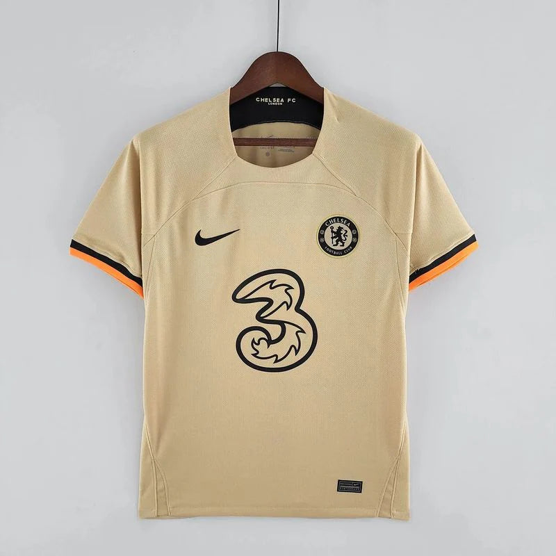 Front view of a pale gold Chelsea FC 2022–2023 short-sleeve third jersey with black chest logos and black and orange sleeve trim.