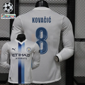 Front and back views of a white Manchester City long-sleeve jersey with blue central stripes, blue Kovačić number 8 print and Etihad Airways sponsor, alongside UCL graphics.