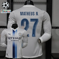 Front and back views of a white Manchester City long-sleeve jersey with blue central stripes, blue Matheus N. number 27 print and Etihad Airways sponsor, alongside UCL graphics.
