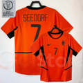 Netherlands orange short-sleeve retro jersey, front and back views with SEEDORF 7 print, black collar and side panels, alongside a 2002 World Cup graphic.
